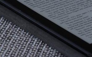 An e-reader device displaying a page of text is partially visible, resting on a textured fabric surface. The focus is on the edge of the device, highlighting its black frame and digital screen with gray text.