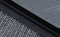 An e-reader device displaying a page of text is partially visible, resting on a textured fabric surface. The focus is on the edge of the device, highlighting its black frame and digital screen with gray text.