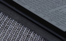 An e-reader device displaying a page of text is partially visible, resting on a textured fabric surface. The focus is on the edge of the device, highlighting its black frame and digital screen with gray text.