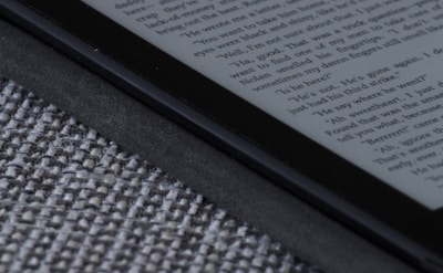 An e-reader device displaying a page of text is partially visible, resting on a textured fabric surface. The focus is on the edge of the device, highlighting its black frame and digital screen with gray text.