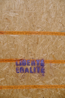 A wooden board with an orange 'Handle with Care' tape running horizontally, displaying text repeated in multiple languages. Stenciled blue letters spell out 'LIBERTÉ EGALITÉ' on the board.