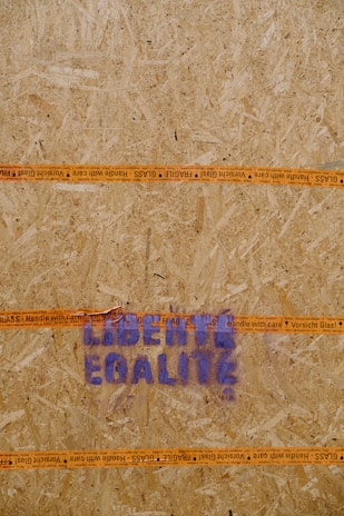 A wooden board with an orange 'Handle with Care' tape running horizontally, displaying text repeated in multiple languages. Stenciled blue letters spell out 'LIBERTÉ EGALITÉ' on the board.