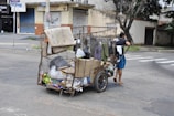 A person is pulling a makeshift cart filled with various items including cardboard boxes and plastic bags, suggesting collection or recycling of materials. The cart is located on a city street with closed shop buildings and some trees in the background.