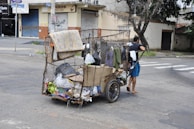 A person is pulling a makeshift cart filled with various items including cardboard boxes and plastic bags, suggesting collection or recycling of materials. The cart is located on a city street with closed shop buildings and some trees in the background.