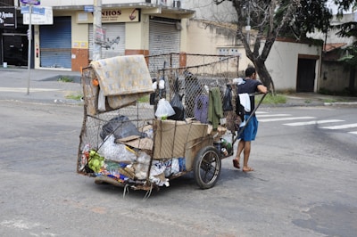 A person is pulling a makeshift cart filled with various items including cardboard boxes and plastic bags, suggesting collection or recycling of materials. The cart is located on a city street with closed shop buildings and some trees in the background.