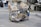 A person is pulling a makeshift cart filled with various items including cardboard boxes and plastic bags, suggesting collection or recycling of materials. The cart is located on a city street with closed shop buildings and some trees in the background.
