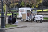 A small ice cream truck is parked in a park, surrounded by green trees and benches. Two chairs are set up next to the truck with a chalkboard menu listing flavors. Two people are sitting on a low wall nearby, engaged in conversation. The scene is calm, with dappled sunlight filtering through the leaves.