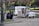 A small ice cream truck is parked in a park, surrounded by green trees and benches. Two chairs are set up next to the truck with a chalkboard menu listing flavors. Two people are sitting on a low wall nearby, engaged in conversation. The scene is calm, with dappled sunlight filtering through the leaves.