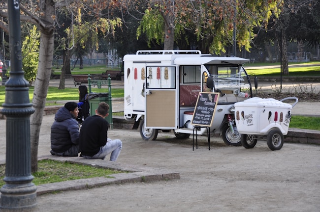 A small ice cream truck is parked in a park, surrounded by green trees and benches. Two chairs are set up next to the truck with a chalkboard menu listing flavors. Two people are sitting on a low wall nearby, engaged in conversation. The scene is calm, with dappled sunlight filtering through the leaves.