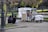 A small ice cream truck is parked in a park, surrounded by green trees and benches. Two chairs are set up next to the truck with a chalkboard menu listing flavors. Two people are sitting on a low wall nearby, engaged in conversation. The scene is calm, with dappled sunlight filtering through the leaves.