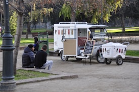A small ice cream truck is parked in a park, surrounded by green trees and benches. Two chairs are set up next to the truck with a chalkboard menu listing flavors. Two people are sitting on a low wall nearby, engaged in conversation. The scene is calm, with dappled sunlight filtering through the leaves.
