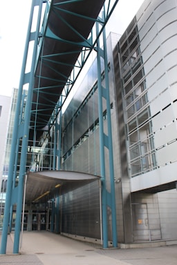 An architectural structure features modern design elements with a prominent walkway bridge supported by blue metal beams. The building exterior is clad in reflective metallic panels, and glass sections offer transparency and a sleek appearance. The structure's overall look is minimalist and industrial, emphasizing straight lines and geometric shapes.