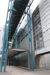 An architectural structure features modern design elements with a prominent walkway bridge supported by blue metal beams. The building exterior is clad in reflective metallic panels, and glass sections offer transparency and a sleek appearance. The structure's overall look is minimalist and industrial, emphasizing straight lines and geometric shapes.