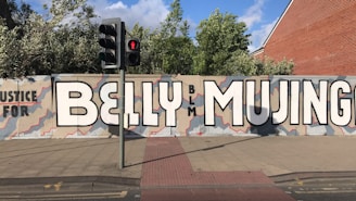 A large mural on a city street wall reads 'Justice for BELLY MUJINGA' with the letters 'BLM' in smaller text. The background is painted with an abstract pattern in shades of beige and gray, with red outlines. Nearby, there is a traffic light showing a red pedestrian signal, and some greenery is visible above the wall.