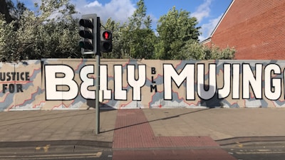A large mural on a city street wall reads 'Justice for BELLY MUJINGA' with the letters 'BLM' in smaller text. The background is painted with an abstract pattern in shades of beige and gray, with red outlines. Nearby, there is a traffic light showing a red pedestrian signal, and some greenery is visible above the wall.
