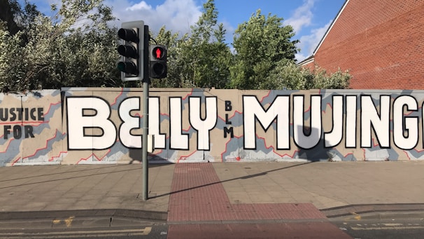 A large mural on a city street wall reads 'Justice for BELLY MUJINGA' with the letters 'BLM' in smaller text. The background is painted with an abstract pattern in shades of beige and gray, with red outlines. Nearby, there is a traffic light showing a red pedestrian signal, and some greenery is visible above the wall.