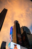 A cityscape featuring tall skyscrapers with bright digital billboards against a glowing orange sky during sunset. The billboards display advertisements for various products and shows.