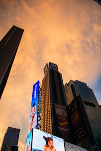 A cityscape featuring tall skyscrapers with bright digital billboards against a glowing orange sky during sunset. The billboards display advertisements for various products and shows.