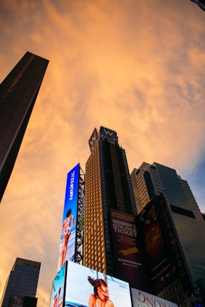 A cityscape featuring tall skyscrapers with bright digital billboards against a glowing orange sky during sunset. The billboards display advertisements for various products and shows.