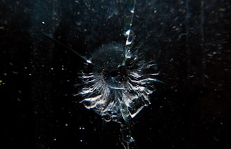 A dark surface with a single point of impact resembling a bullet hole, surrounded by radiating cracks and fragments of glass, highlighting concentric patterns.