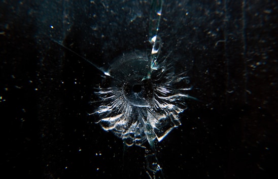 A dark surface with a single point of impact resembling a bullet hole, surrounded by radiating cracks and fragments of glass, highlighting concentric patterns.