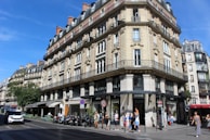 A classic Parisian street scene featuring an ornate, multi-story building with intricate stonework and wrought iron railings. The ground floor contains several shops, including a Guess store. People are walking along the sidewalk, and a few vehicles are moving along the street. The sky is clear and blue, suggesting a sunny day.