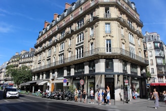 A classic Parisian street scene featuring an ornate, multi-story building with intricate stonework and wrought iron railings. The ground floor contains several shops, including a Guess store. People are walking along the sidewalk, and a few vehicles are moving along the street. The sky is clear and blue, suggesting a sunny day.