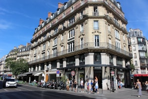 A classic Parisian street scene featuring an ornate, multi-story building with intricate stonework and wrought iron railings. The ground floor contains several shops, including a Guess store. People are walking along the sidewalk, and a few vehicles are moving along the street. The sky is clear and blue, suggesting a sunny day.