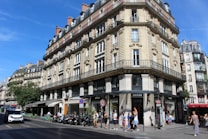 A classic Parisian street scene featuring an ornate, multi-story building with intricate stonework and wrought iron railings. The ground floor contains several shops, including a Guess store. People are walking along the sidewalk, and a few vehicles are moving along the street. The sky is clear and blue, suggesting a sunny day.
