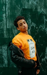 A person poses against a textured, dark green wall wearing a bright orange t-shirt with a 'Skim Milk' graphic and a partially worn black leather jacket. The person gazes directly at the camera, exuding confidence.