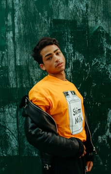 A person poses against a textured, dark green wall wearing a bright orange t-shirt with a 'Skim Milk' graphic and a partially worn black leather jacket. The person gazes directly at the camera, exuding confidence.