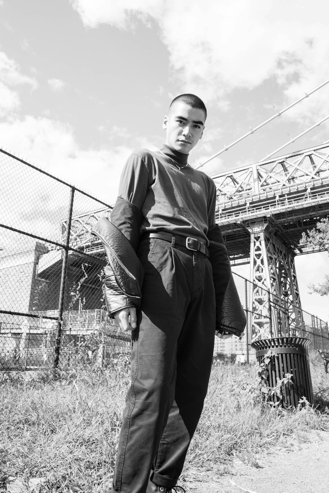 grayscale photo of man in jacket and pants standing near metal fence