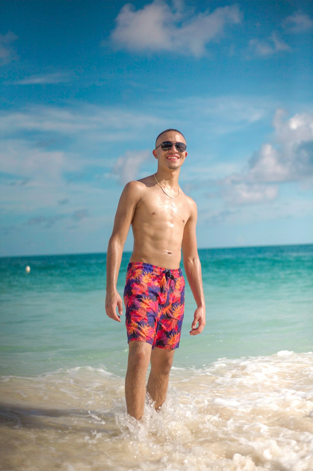 man in blue and red floral shorts wearing black sunglasses standing on beach during daytime