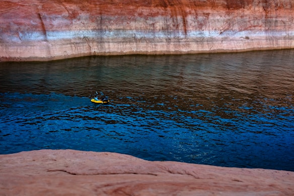 A person in a small yellow kayak paddles on a calm body of water that reflects the surrounding reddish-brown rock formations. The water has a vibrant blue hue, contrasting with the earthy tones of the canyon walls.
