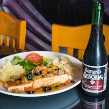 A plate of food containing a grilled piece of fish topped with olives, celery, and assorted vegetables, accompanied by mashed potatoes and rice. A salad with lettuce and a slice of tomato is also on the plate. Next to the plate, there is a bottle of Sangría Señorial, a non-alcoholic beverage. Two wooden chairs are in the background.