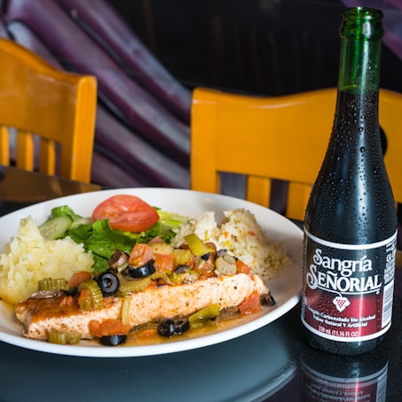 A plate of food containing a grilled piece of fish topped with olives, celery, and assorted vegetables, accompanied by mashed potatoes and rice. A salad with lettuce and a slice of tomato is also on the plate. Next to the plate, there is a bottle of Sangr&iacute;a Se&ntilde;orial, a non-alcoholic beverage. Two wooden chairs are in the background.