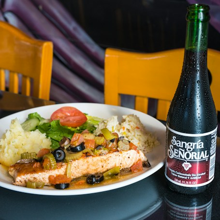 A plate of food containing a grilled piece of fish topped with olives, celery, and assorted vegetables, accompanied by mashed potatoes and rice. A salad with lettuce and a slice of tomato is also on the plate. Next to the plate, there is a bottle of Sangría Señorial, a non-alcoholic beverage. Two wooden chairs are in the background.