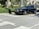 A luxurious black convertible car is parked on a sunlit street, surrounded by lush greenery and upscale buildings.
