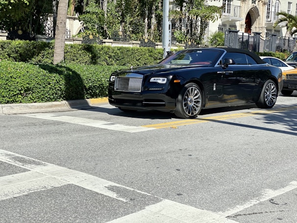 A luxurious black convertible car is parked on a sunlit street, surrounded by lush greenery and upscale buildings.