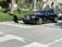 A luxurious black convertible car is parked on a sunlit street, surrounded by lush greenery and upscale buildings.