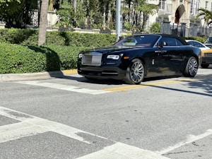 A luxurious black convertible car is parked on a sunlit street, surrounded by lush greenery and upscale buildings.