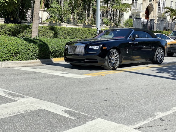 A luxurious black convertible car is parked on a sunlit street, surrounded by lush greenery and upscale buildings.