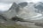 A remote mountain cabin is perched on a rugged hillside surrounded by steep, rocky terrain. In the background, expansive snow-covered peaks and a large glacier exhibit varying shades of gray and white under a cloudy sky.