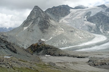 A remote mountain cabin is perched on a rugged hillside surrounded by steep, rocky terrain. In the background, expansive snow-covered peaks and a large glacier exhibit varying shades of gray and white under a cloudy sky.