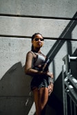 A person stands against a concrete wall, wearing sunglasses and a mesh top. The lighting creates strong shadows, emphasizing the textures of the wall and the outfit.