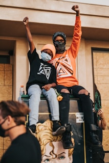 Two people are sitting on top of a structure, raising their fists as a sign of solidarity or protest. They are wearing graphic shirts and masks. A man is walking by with a mask, and a colorful artwork is visible on the structure they are sitting on. One person holds a flower, and there is a sign partially visible in the background.