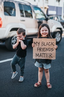 Two young children are standing on a street, with a large vehicle parked in the background. The child on the right is smiling and holding a cardboard sign that says 'MY BLACK FRIENDS MATTER.' The child on the left appears to be giggling or shy, with both hands near their face.