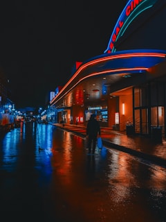 A nighttime street scene featuring a brightly lit cinema with neon lights. The wet pavement reflects the vivid blues and reds from the signage. A solitary figure walks down the street carrying a bag.
