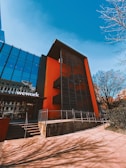 A modern building with a glass facade and a bold orange accent wall stands against a clear blue sky. The structure features black metal louvers on the orange part, stairs leading to the entrance, and a sign with the word 'wework' on the glass section. The surrounding area includes bare tree branches, indicating a possibly urban setting.