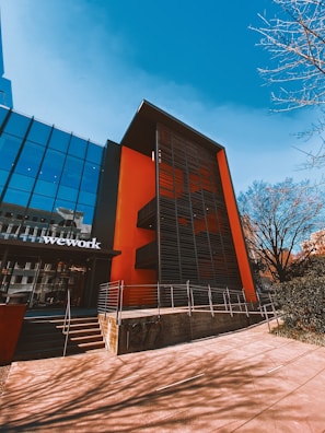 A modern building with a glass facade and a bold orange accent wall stands against a clear blue sky. The structure features black metal louvers on the orange part, stairs leading to the entrance, and a sign with the word 'wework' on the glass section. The surrounding area includes bare tree branches, indicating a possibly urban setting.